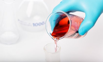 Laborbecher mit roter Flüssigkeit wird von Hand mit Handschuh in Glasgefäß gegossen – Laboranalyse